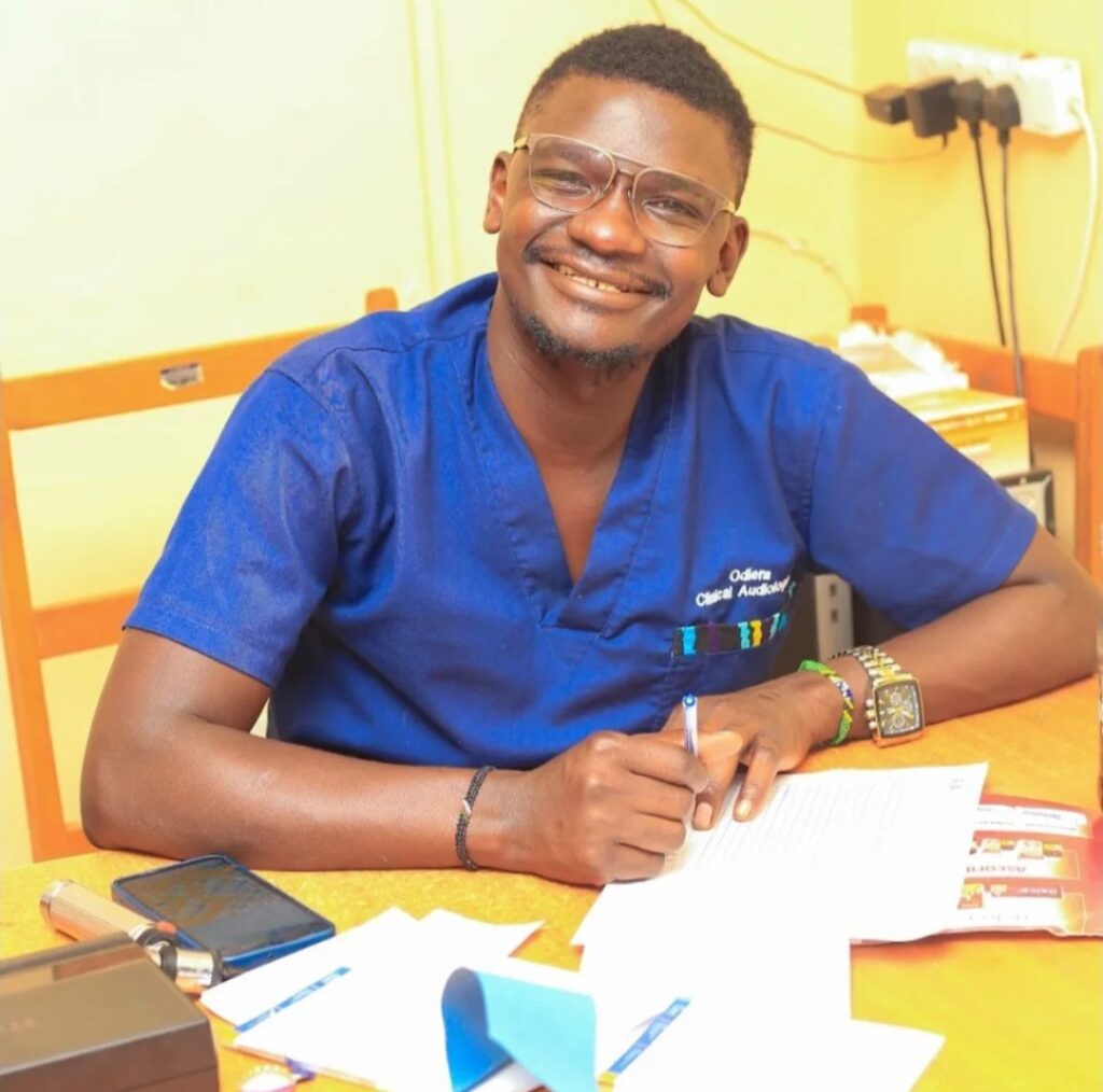 Audioloog Okoth uit Siaya, Kenia, glimlacht terwijl hij werkt aan documenten voor gehoorprojecten met steun van WeHearU